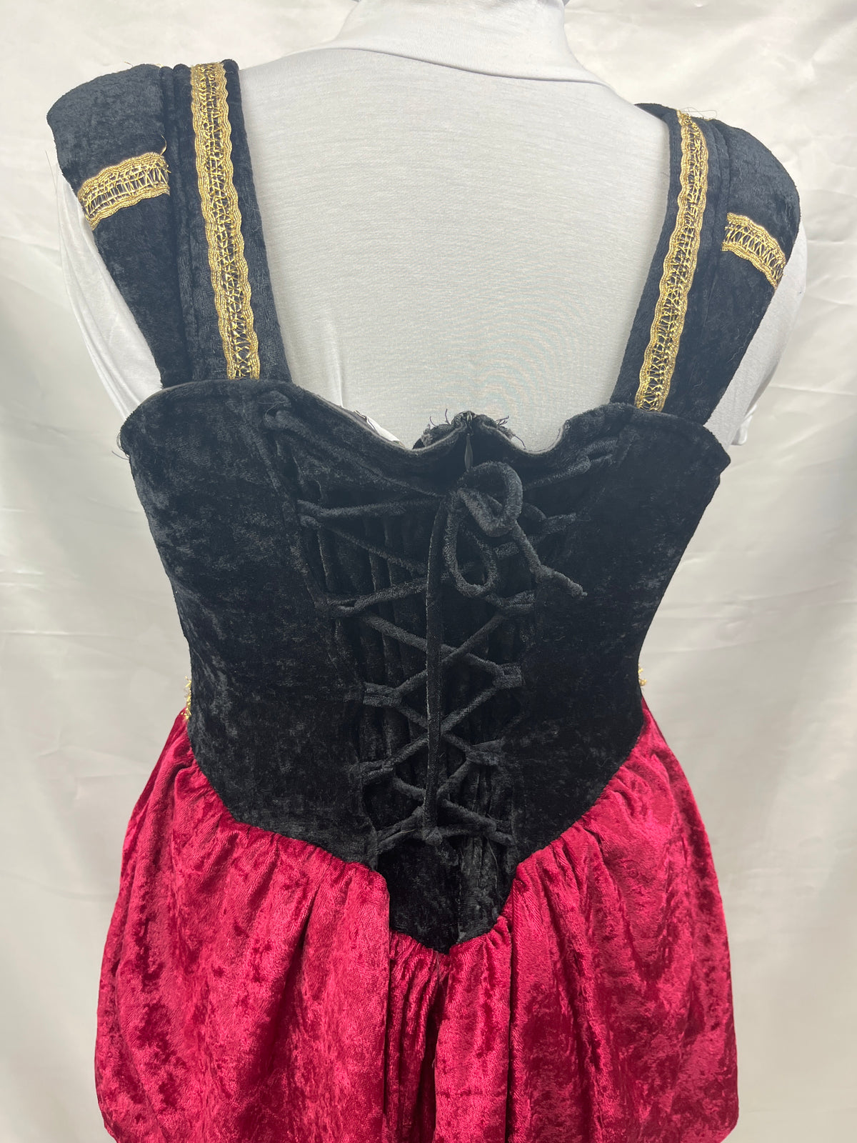 A medieval-style dirndl dress displayed on a white mannequin torso, showing the back view. The dress features a black velvet lace-up corset bodice with wide shoulder straps trimmed in gold embroidery, and a bright pink velvet skirt.